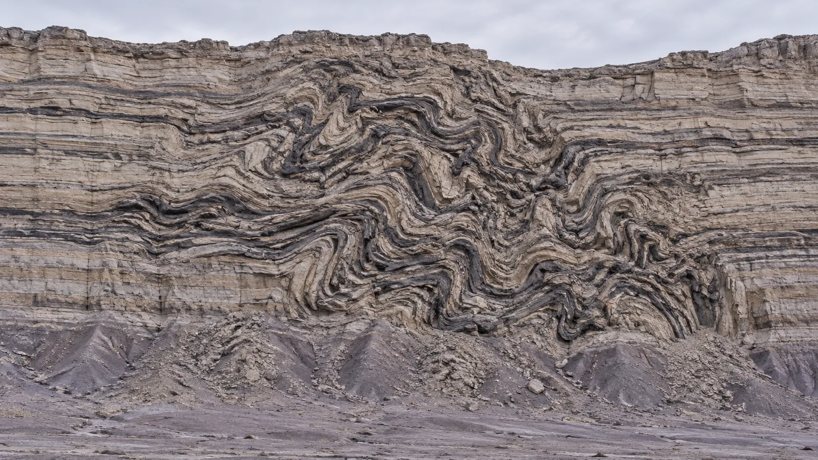A grey badlands cliff is neatly layered at the top and bottom, and in the middle the strata twist and loop into a knot. A few million years went sideways.